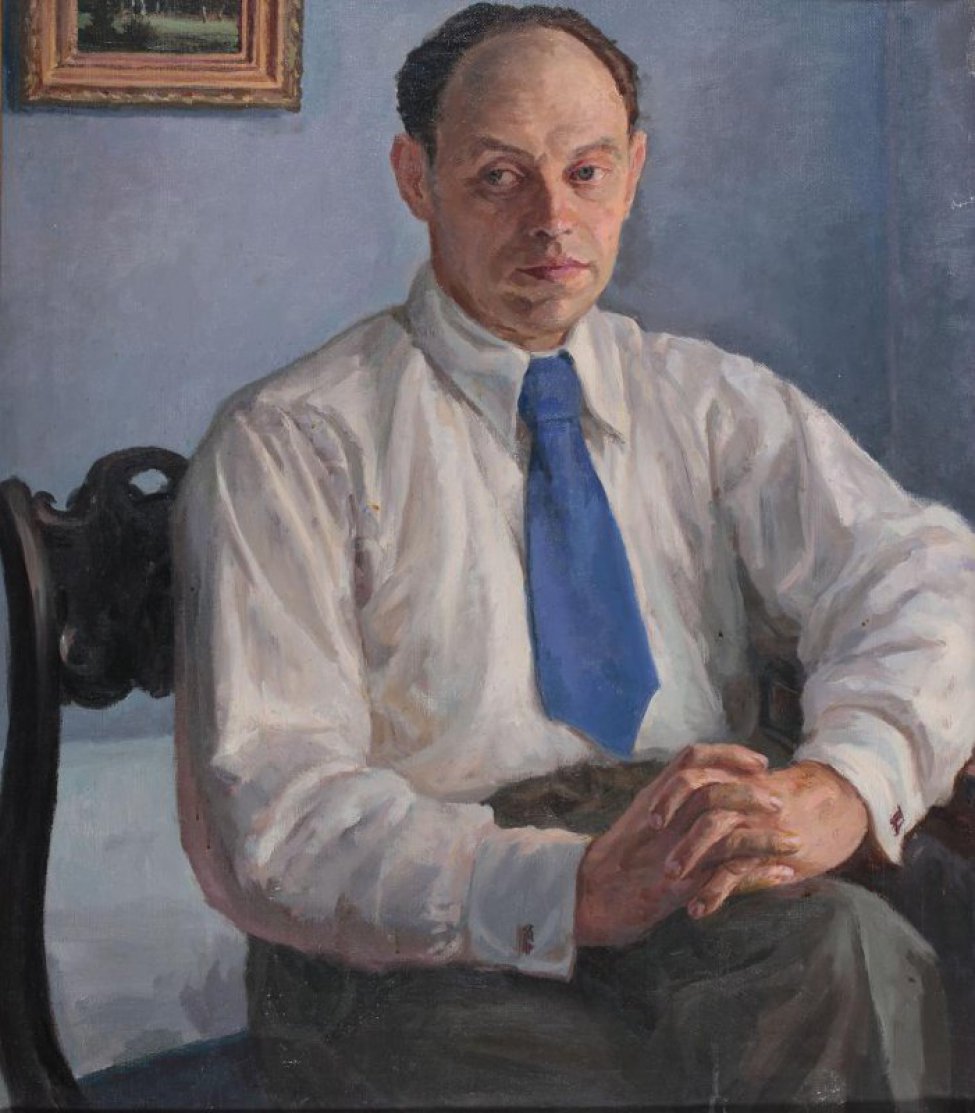 На светло-голубом фоне поколенное изображение мужчины средних лет, сидящего на стуле, в белой рубашке с синим галстуком.