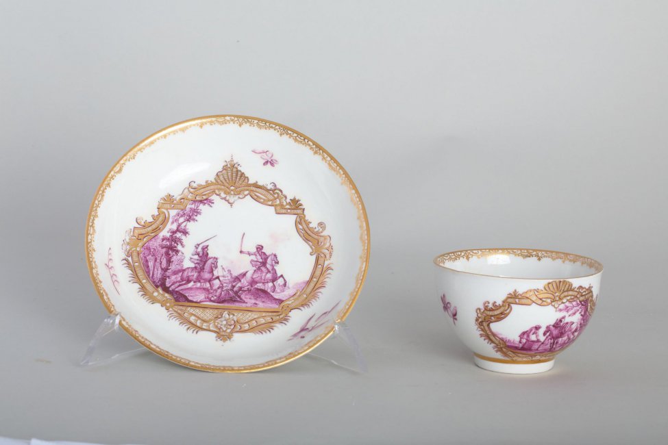 Чашка в форме боль, белая, по борту декорирована двумя батальными композициями и цветами малинового (пурпурового) цвета; на дне чашки - малиновый цветок; по внутреннему краю - орнаментальная золоченая полоска.