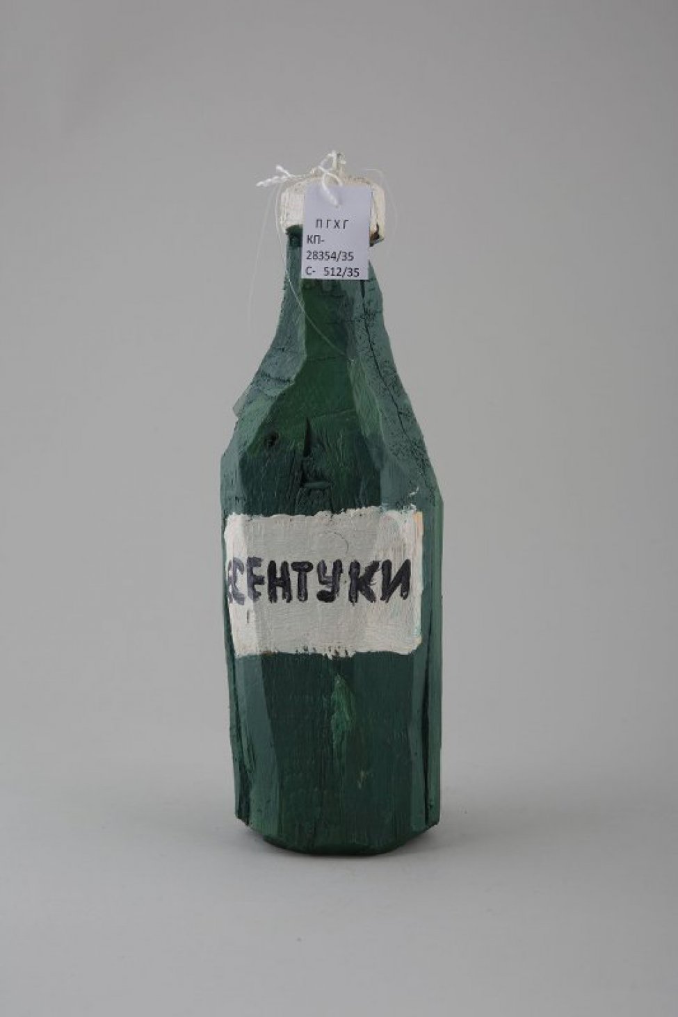 Стилизованное изображение бутылки темно-зеленого цвета, с белой крышкой и белой этикеткой по центру бутылки с надписью черной краской: ЕСЕНТУКИ.  В центре крышки металлическое колечко.