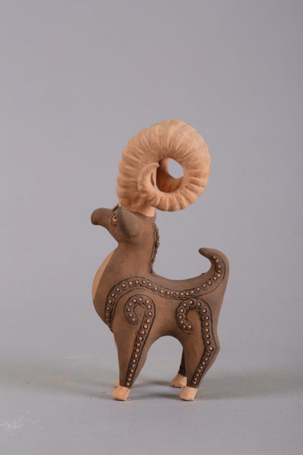 Стилизованная фигура барана с высокими ребристыми, закручивающимися  рогами; ноги, уши, лоб и спина декорированы коричневыми поясками из чередующихся, рельефных шариков бежевого цвета