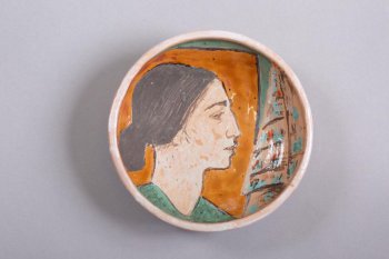 Блюдо круглое, с пологим высоким бортом, на зеркале изображен профиль темноволосой женщины (волосы гладко зачесаны и убраны в пучок) на абстрактном геометрическом фоне.