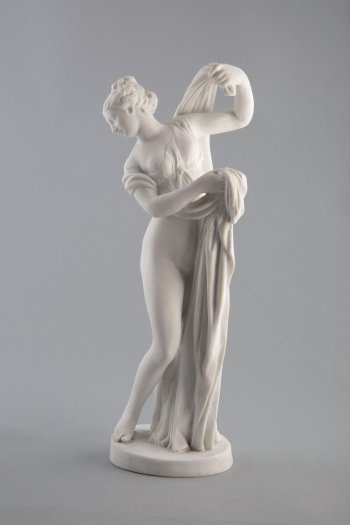 Фигура полуобнаженной женщины  на круглом постаменте. По левой половине ее тела с плеч до пола спускается складками одежда, края которой она поддерживает руками. Волосы женщины собраны в прическу.