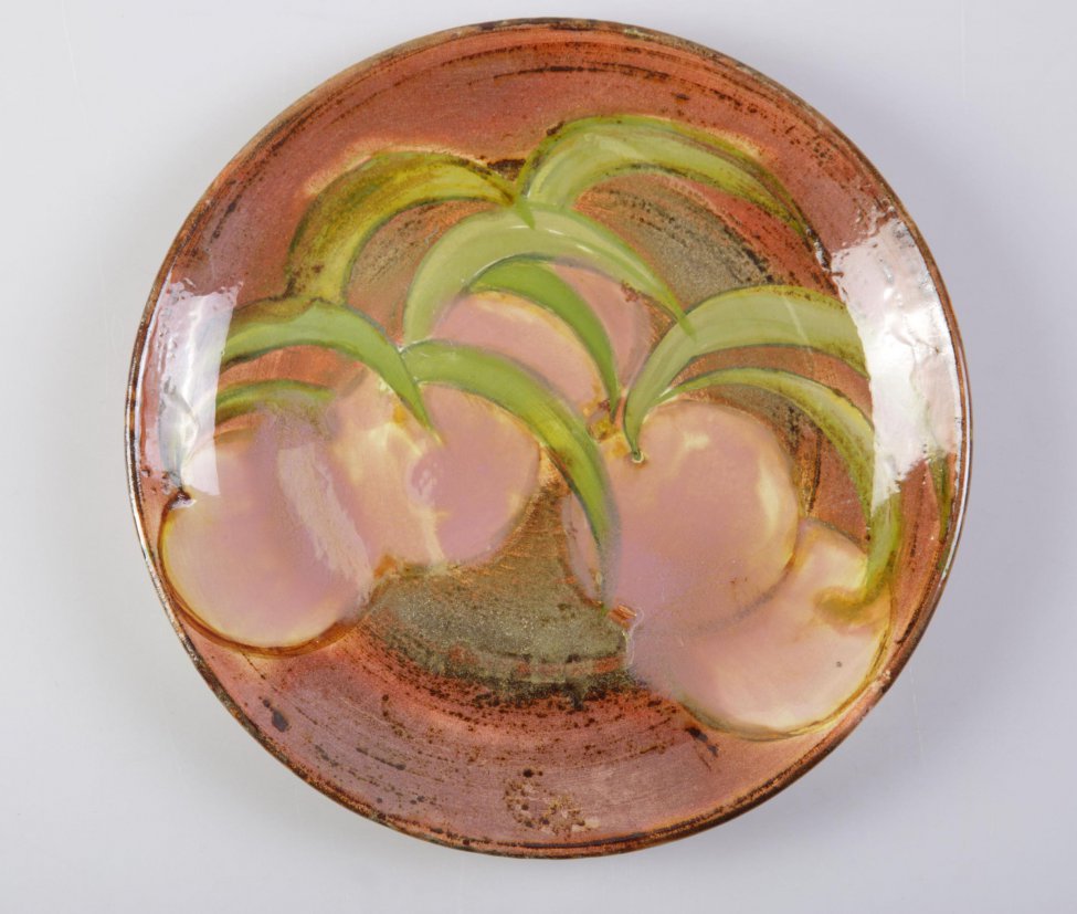 Тарелка круглая, плоская. Покрыта поливой розового цвета. На дне тарелки пять розово-малиновых плодов (персиков) и выходящие из них зеленые полукружия листьев (9 штук). На обратной стороне тарелки по белому фону проступают следы коричнево-розовой поливы.