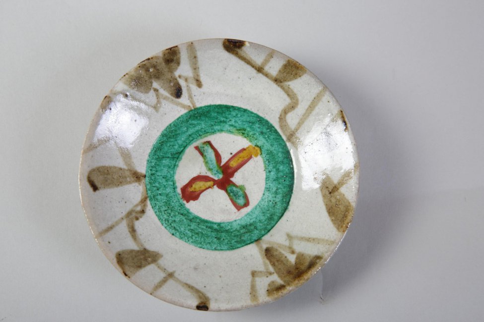 Блюдце на низкой ножке, серо-белого цвета. Украшено зелёным кольцом с красным крестом внутри (на дне) и коричневыми разводами по борту.