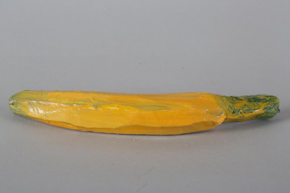 Обобщенное изображение банана желтого цвета с зелеными разводами, с длинной желто-зеленой веткой, к которой прикреплена металлическая петелька.