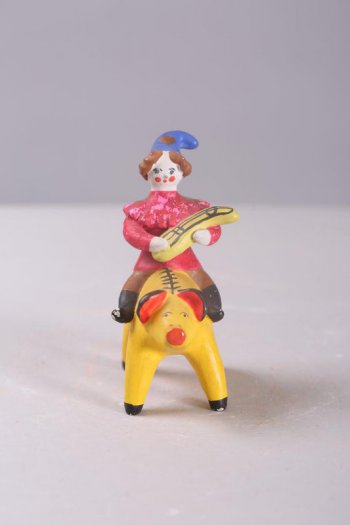 Клоун сидит на желтой свинье верхом. Он в красном кафтане, остроконечной синей шляпе и черных сапогах. В руках у него желтый музыкальный инструмент. На его шляпе и плечах, а так же на боках свиньи - позолота.