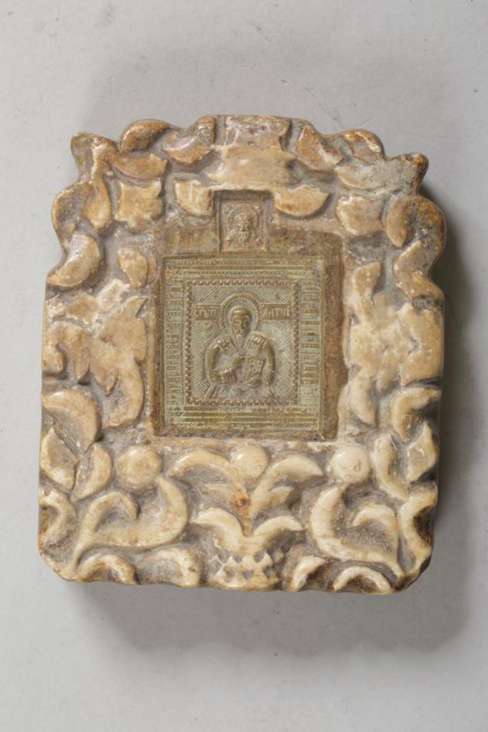 Литая икона в оправе из резного камня, с навершием "Спас нерукотворный". В центре изображен святой с благословляющим жестом правой руки и Евангелием в левой руке.