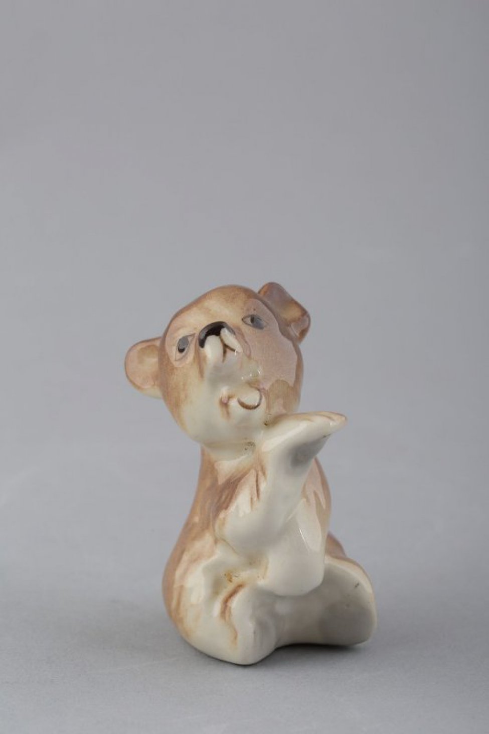 Фигурка сидящего медвежонка с открытой пастью и  поднятой вверх правой лапой, левая - опущена вниз; роспись: медвежонка бежево-коричневым цветом; глаза и нос - темно-коричневым.