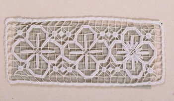 Образец в виде ажурной полосы из белых нитей. На тонкой  сетке в одну нить иглой шит рельефный узор из трех четырехлепестковых розеток. Образец пришит к картонному листу.