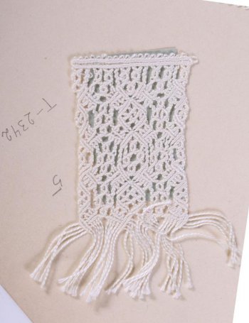 Образец белого цвета в виде широкой полосы со сложным плетёным узором из ромбов и небольших кружков. На одном конце свободно висящие нити. Образец пришит к картонному листу.