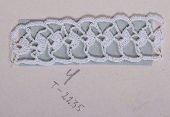 Образец белого цвета в виде узкой ажурной полосы, по центру которой проходит вертикальный ряд из веерообразных фигур. Образец пришит к картонному листу.