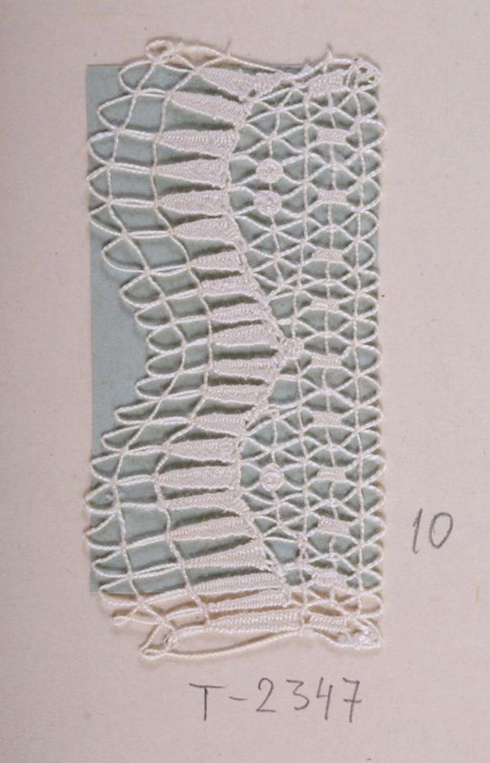Образец белого цвета в виде ажурной полосы, один край которой составлен двумя фестонами. По образцу проходит волнистая линия, повторяющая фестоны из плотно сплетённых зубчатых столбиков. Образец пришит к картонному листу.