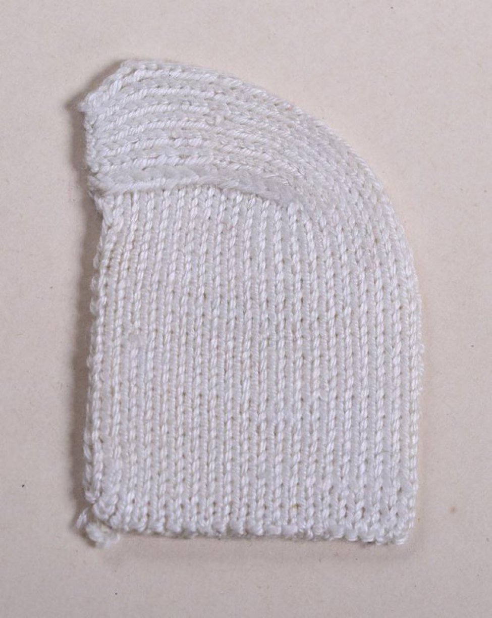 Образец прямоугольной, закругленной с одного угла формы из белых нитей  выполнен в технике вязки лицевыми петлями. Образец демонстрирует вязание пятки чулка или носка.