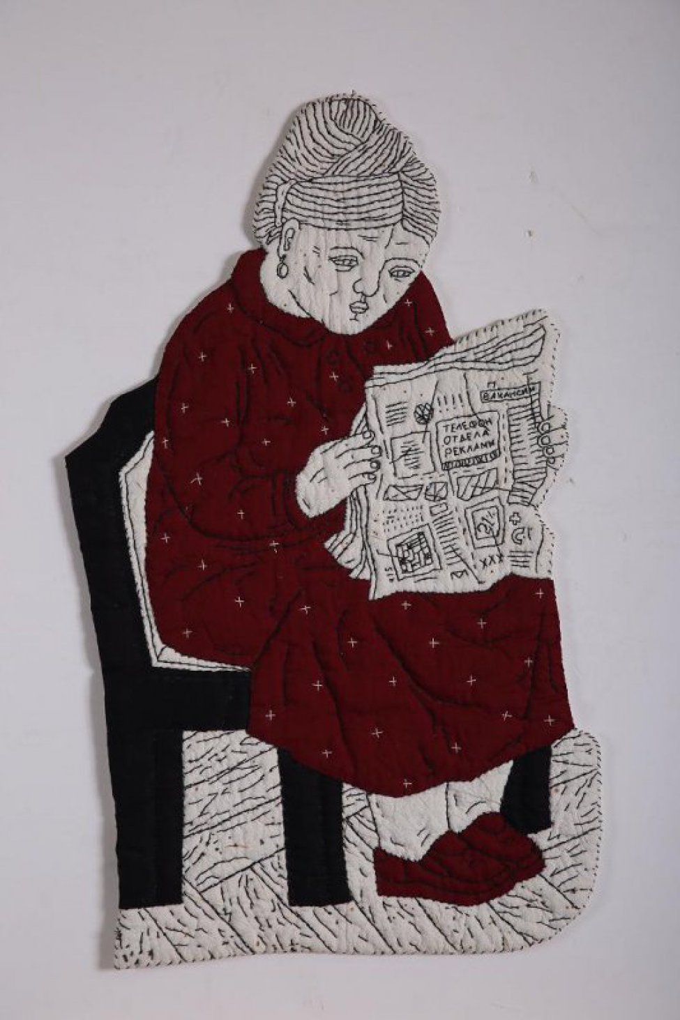 По абрису вырезана сидящая женская фигура в темно-красном платье, с газетой в руках, на черном стуле.  Рисунок фигуры  вышит черными нитями.  Женщина изображена вправо, с высокой прической, с овальной серьгой в ухе, в темно-красных туфлях, платье расшито отдельными белыми крестиками, в тексте газеты слова: "ВАКАНСИИ", "ТЕЛЕФОН | ОТДЕЛА | РЕКЛАМЫ",  "21", "415", МОНОГРАММА "ПМ".  Местами по поверхности белой ткани небольшие пятна и полоски прожига. Вся фигура по краю обшита черной нитью. Внутри всей фигуры слой ватина. Оборот подложен белой бязью и простеган черными нитями.  С оборота в верхней части пришиты 2  плоских стальных кольца.