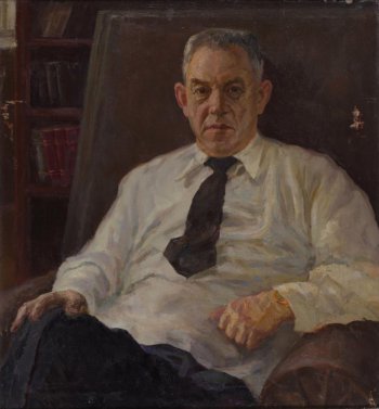 На фоне стеллажа с книгами дано поколенное изображение пожилого сидящего человека в светлой рубашке с темным галстуком, в темных брюках.