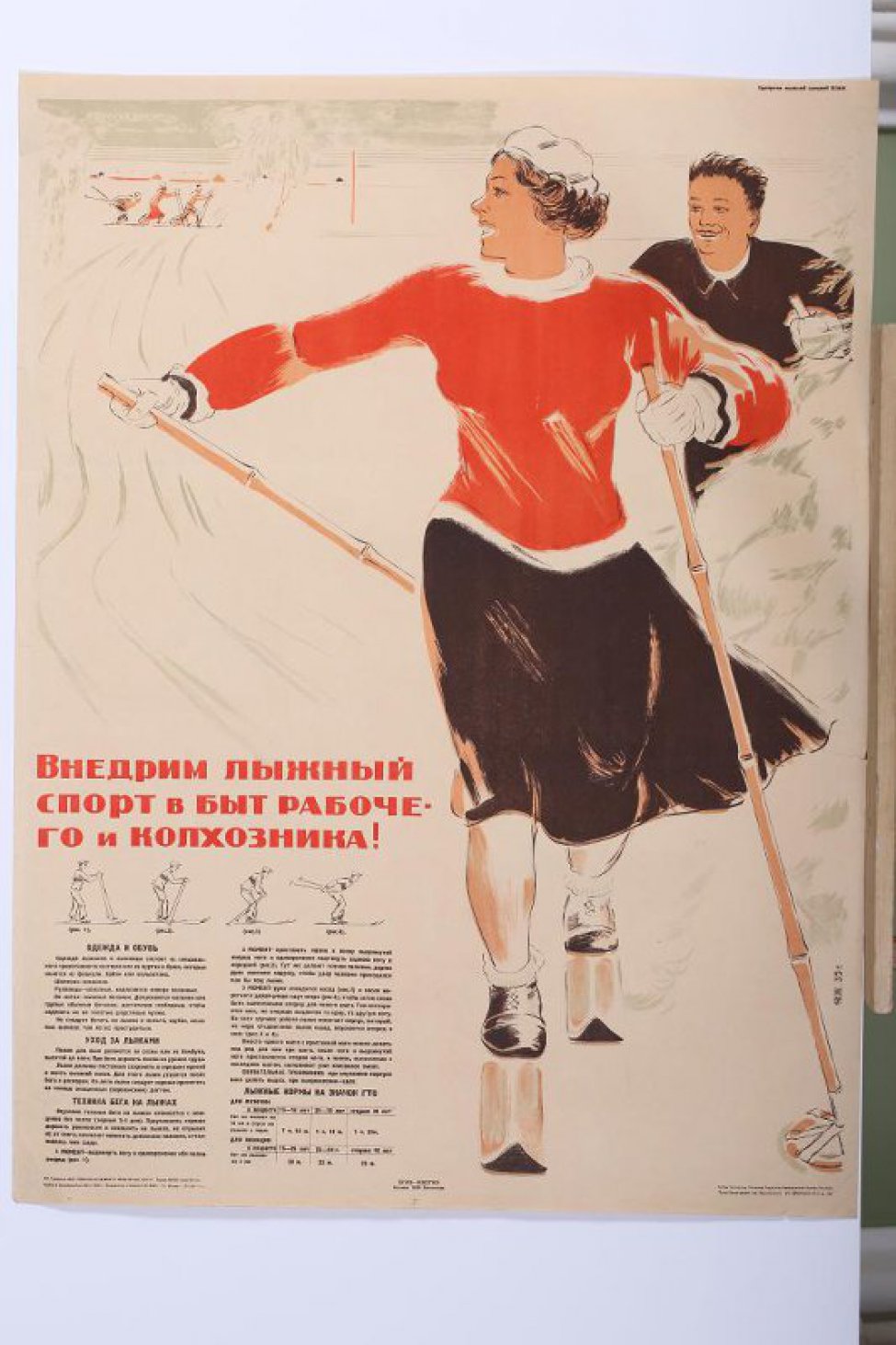 Изображена на зимнем пейзаже молодая лыжница в черной юбке, красном свитере и белом берете. В руках лыжные палки. За ней мужчина лыжник без шапки. Внизу слева текст:" Одежда... вдох".