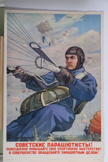 Изображен прицелелившийся парашютист  в синем комбинезоне и шлеме. За ним в небе и на земле-еще парашютисты..