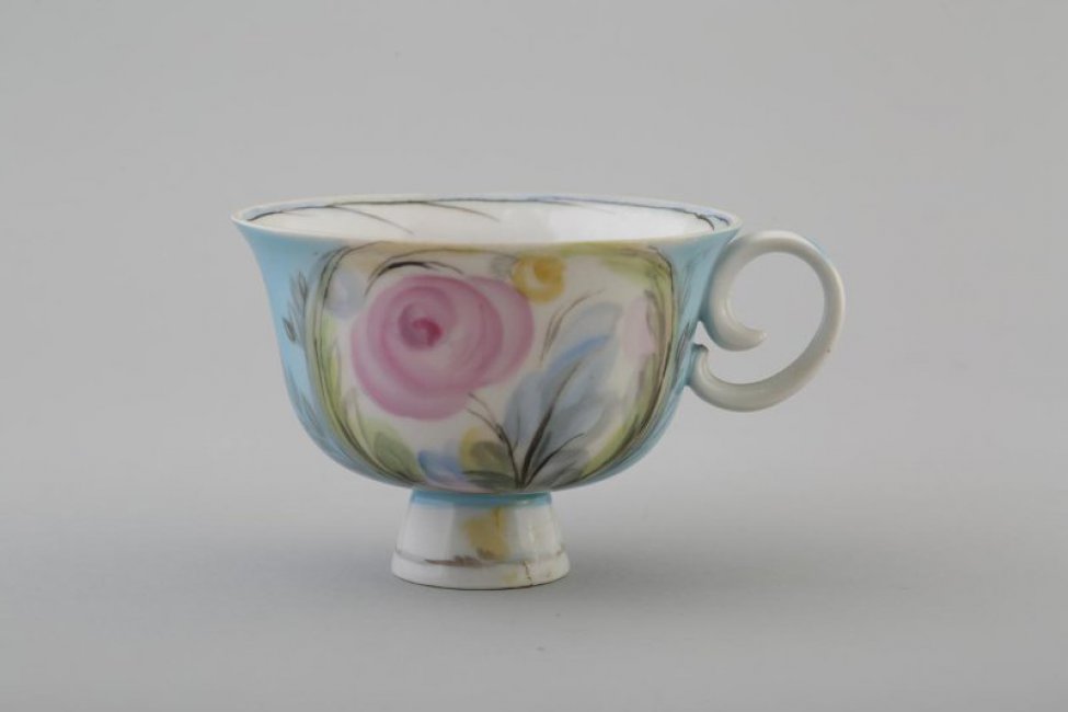 чашка вазообразной формы с расширяющимся кверху  туловом, на высокой круглой ножке, с ручкой. На белом фоне стилизованное  изображение розы в окружении листьев.