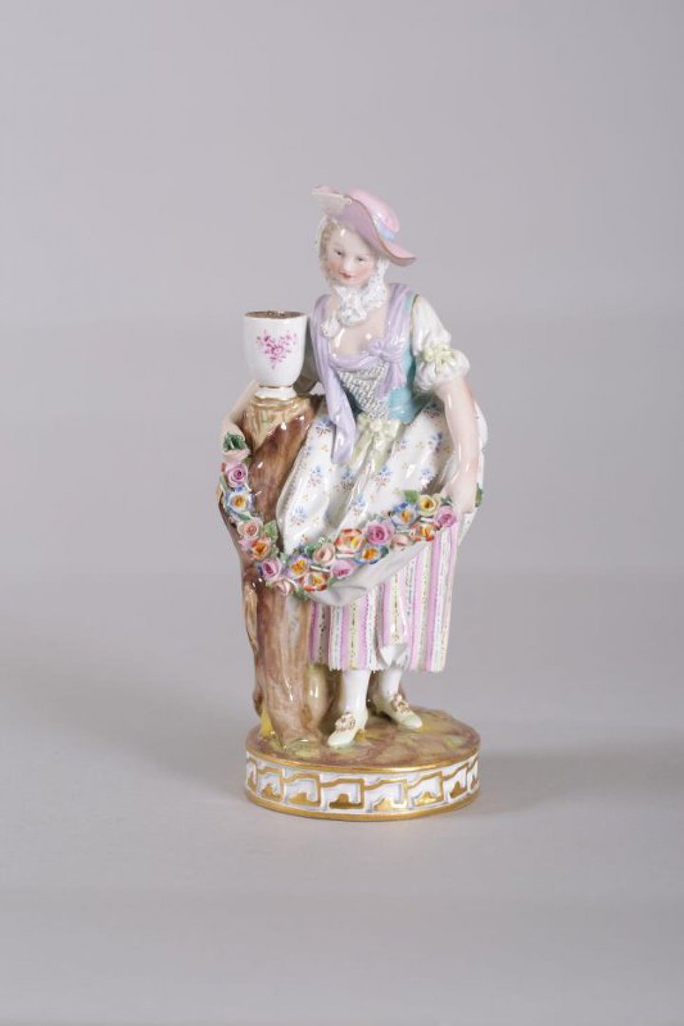Фигура на круглом постаменте. Изображена молодая женщина в розовой шляпке с голубой лентой. Одной рукой она обняла ствол дерева с вазой наверху, в руках гирлянда цветов. Кругом по боковой стороне постамента золоченый орнамент.