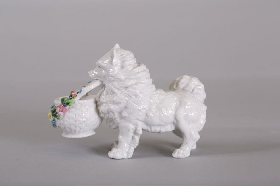 Фигурка лохматой белой собаки с корзинкой в зубах. По борту корзинки лепная расписаная гирлянда цветов.