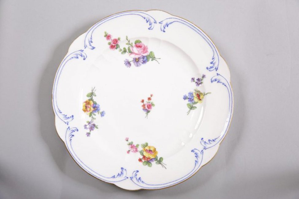 Тарелка мелкая, белая, с волнистым золоченым краем. Борт декорирован рельефными синими рокайльными завитками; дно расписано пятью цветочными букетами с розовыми и желтоватыми розами.
