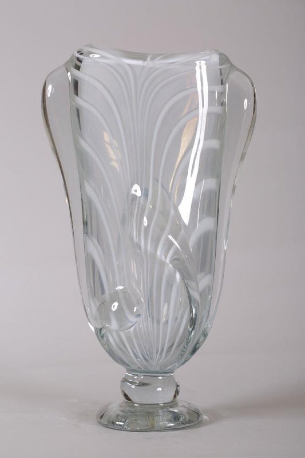 ваза серого цвета, плоская, с белыми полосами из толстого стекла; форма вытянутая, расширяющейся кверху, на ножке. По тулову - налепы, два из которых имитируют ручки. Налепы и ножка из прозрачного стекла.