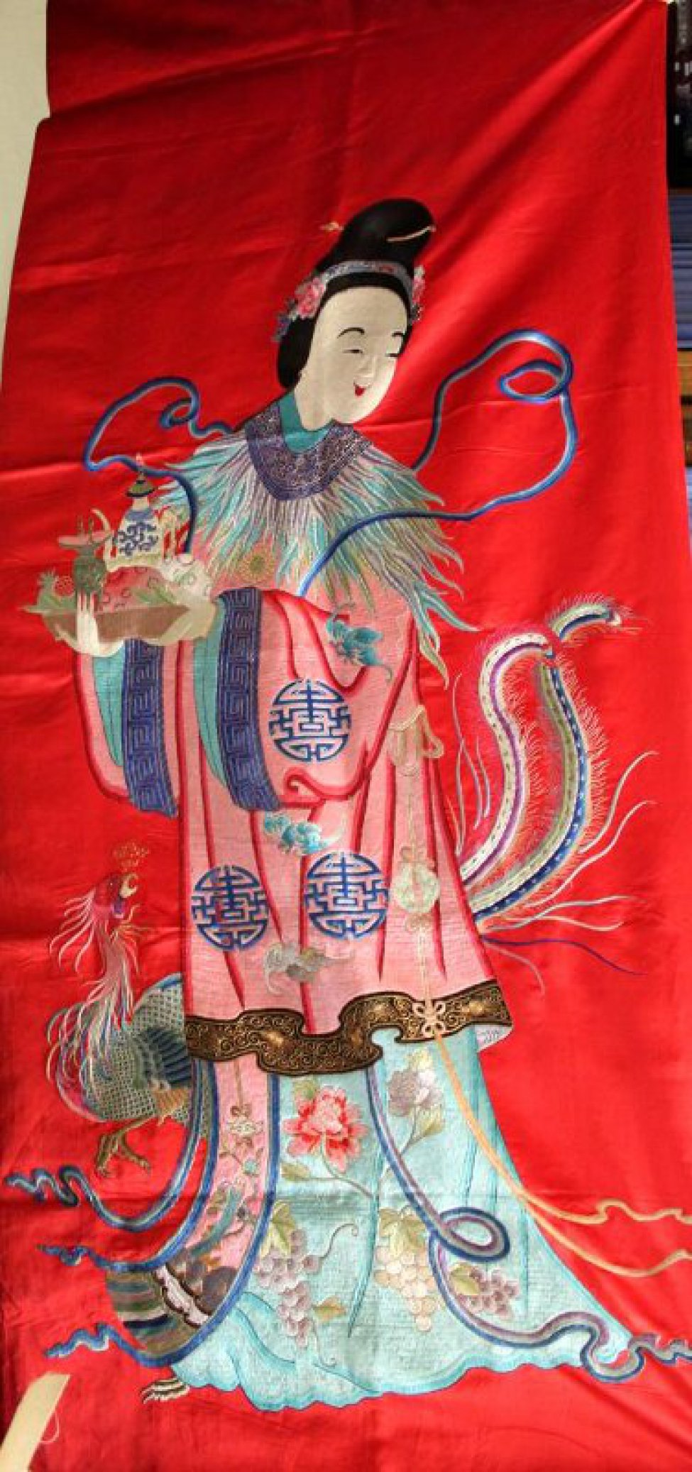 Вертикальной прямоугольной формы из красного атласа с вышитой разноцветными шелками фигурой женского божества. Изображена женщина с высокой прической темно-коричневых волос, украшенной сине-голубой диадемой с цветком по бокам. В верхней части прически - золотая булавка. В руках ее - влево - поднос с тремя фруктами (два персика и гранат), белым чайником с  синим орнаментом и зеленым сосудом на высоких ножках. Верхнее платье розового цвета с черной каймой внизу. Узор - три крупных синих иероглифа и три летучие мыши. По краю широких рукавов синяя кайма. Ворот в виде широкой синей каймы с золотым рисунком и со спускающимися вниз разноцветными перьями. Нижняя одежда голубого цвета с двумя пионами и гроздьями винограда. Внизу, за фигурой, изображена птица феникс (слева выступают ее голова, часть туловища и одна нога), справа - два длинных тонких пера. Около плеч фигуры - длинная тонкая сине-голубая лента. Слева внизу из-под платья выступает часть птичьей лапы. Детальные разработки рисунка вышивки выполнены золотными нитями.